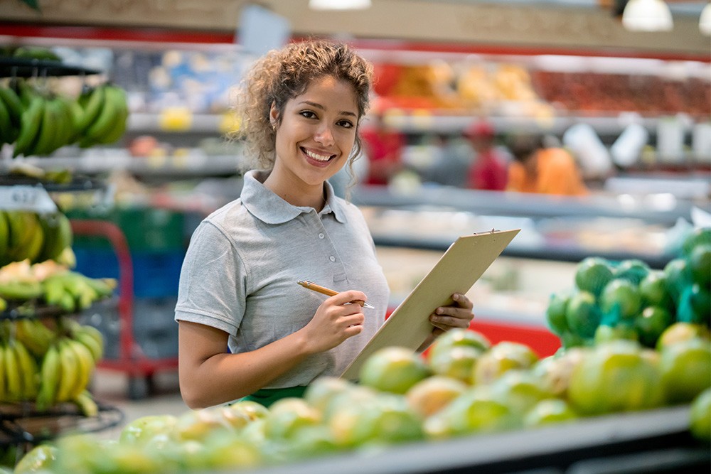A young woman working at a food retail grocery store holds a clipboard in front of fruits and vegetables.