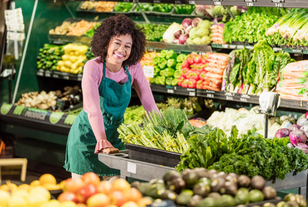 A young female food retail employee works at a grocery store pushing a cart full of vegetables.