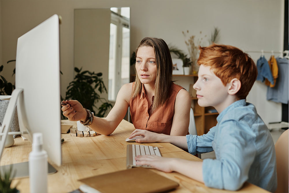 A young boy works on the computer with his Mom while she teaches him.