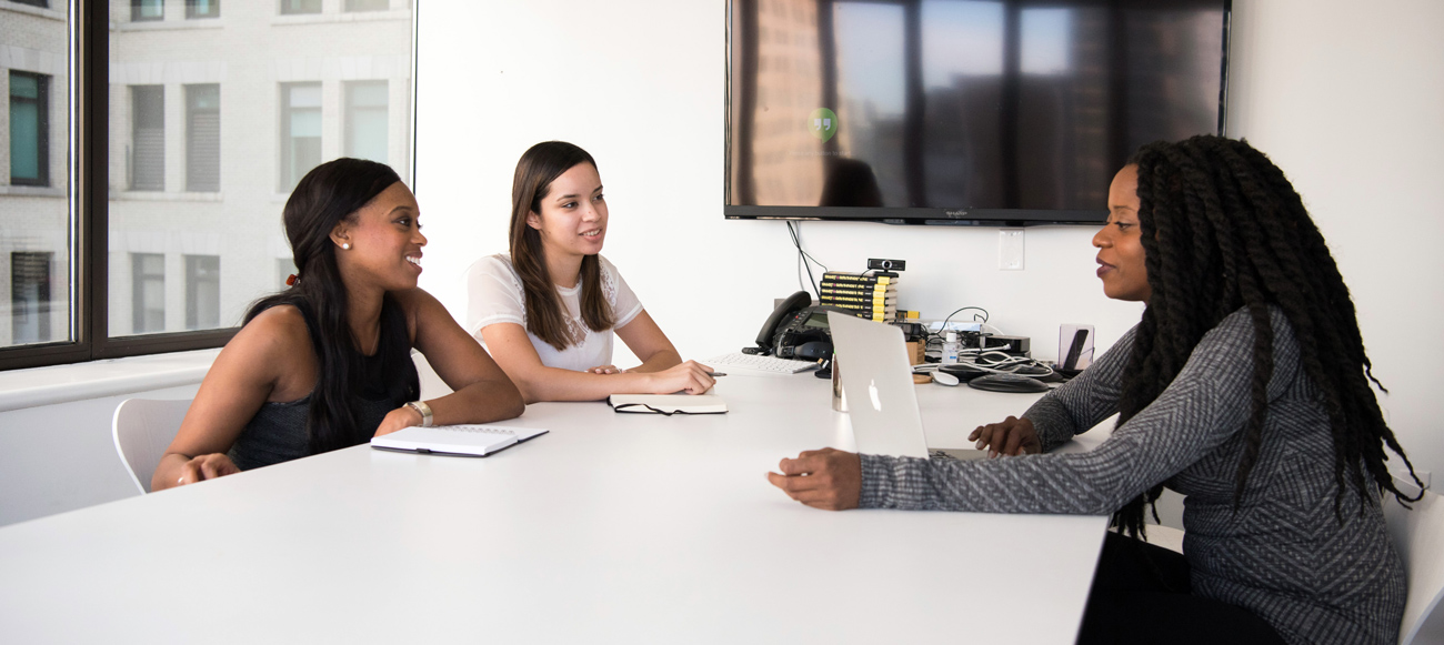 three women sit at a conference table, speaking with each other