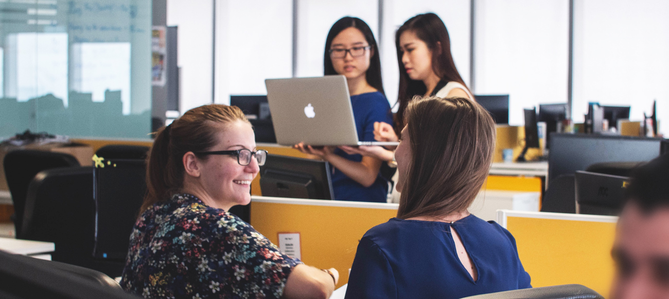 coworkers talk at their desks while two other coworkers walk by in the background