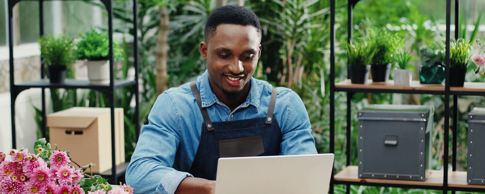 Photo: a smiling young man works at a laptop in his floral shop