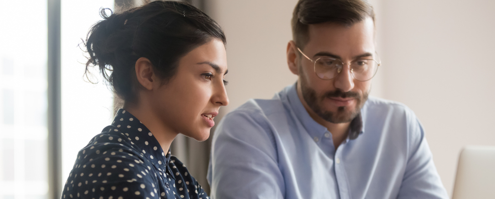Photo: A female employee explains the purpose of a learning management system to a male employee as they both look at a laptop.