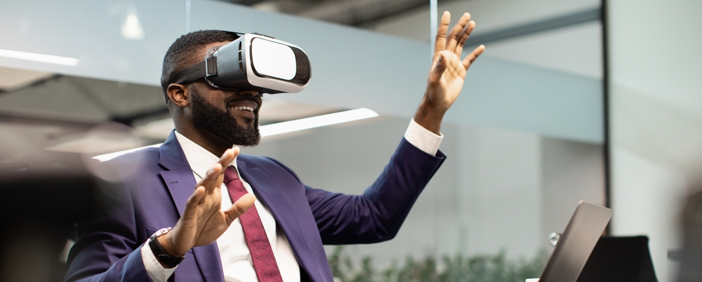 Photo: A businessman sits at his desk using a VR headset, he is excited to try the new technology.
