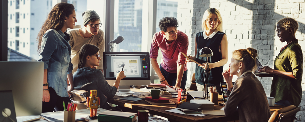 Photo: A diverse group of employees surrounding an office desk work together to plan a project.