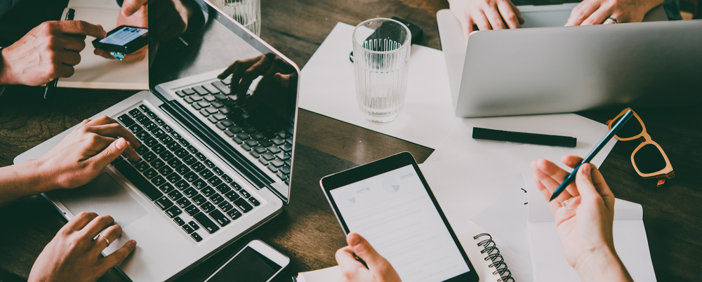 Photo: People work together around a table on laptop computers, smartphones, and tablets. Only their hands, the devices, and the top of the table are shown.