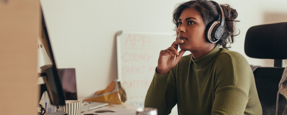 Photo: A young woman wearing headphones looks away from her laptop, deep in thought.