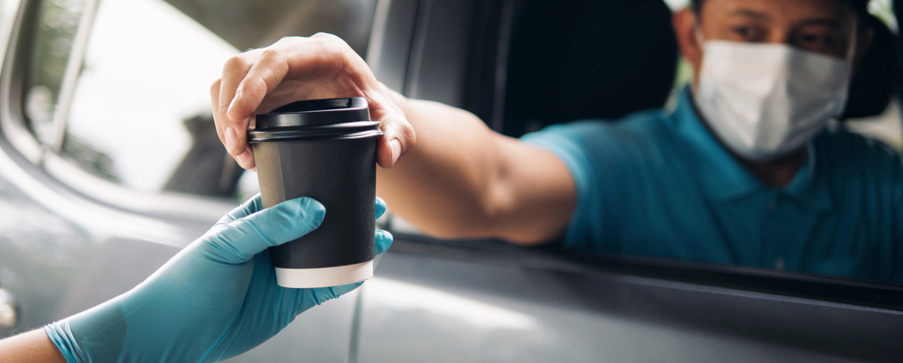 Drive thru food and drink service during Coronavirus COVID-19 pandemic. Staff hand in protective glove serving a coffee cup to customer a car driver man in protective face mask at drive thru service.