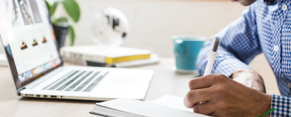 person writing on notebook in front of laptop