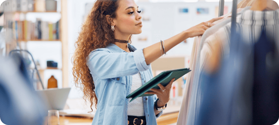 A person with curly hair is using a tablet while selecting clothes on a clothing rack in a retail store.
