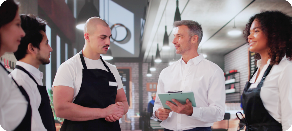 A group of four employees in a restaurant setting, dressed in uniforms, engaged in discussion with a man holding a clipboard.

