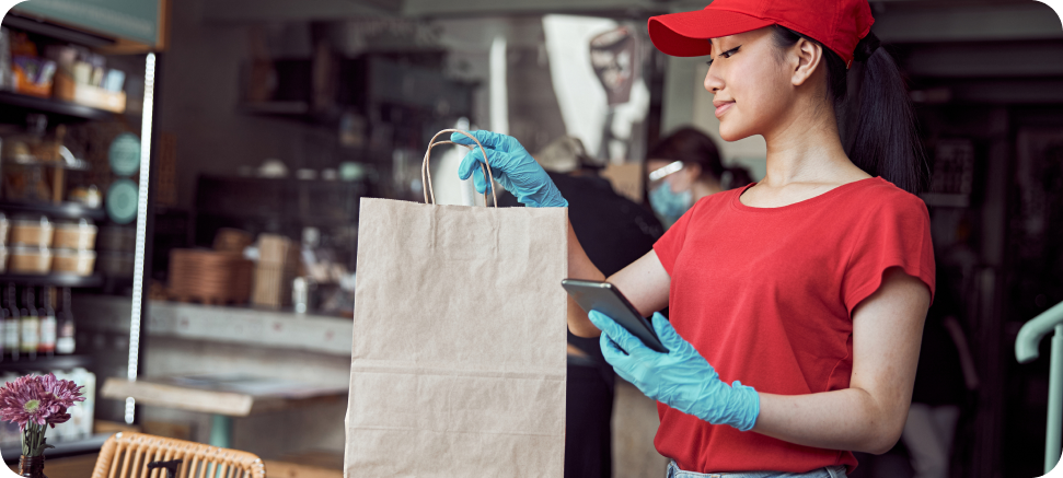 A woman in a red shirt and cap holds a brown paper bag while looking at her phone, wearing blue gloves, in a café setting.