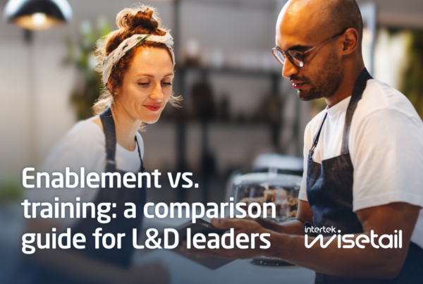 Two people in aprons are talking in a warmly lit, blurred kitchen setting. Text reads, "Enablement vs. training: a comparison guide for L&D leaders." Mood is collaborative.
